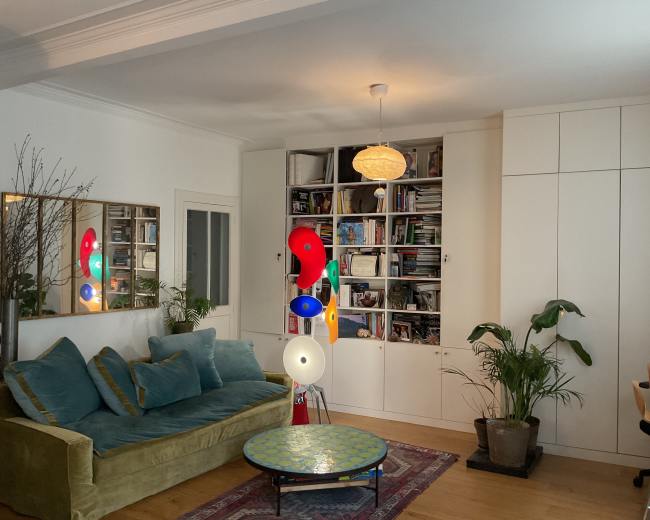 The image shows a cozy living room with a green sofa, a decorative round table, bookshelves filled with items, and several indoor plants.