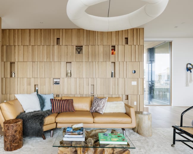 A modern living room features a light brown leather sofa, a glass coffee table, and a wooden accent wall with various decorative items.