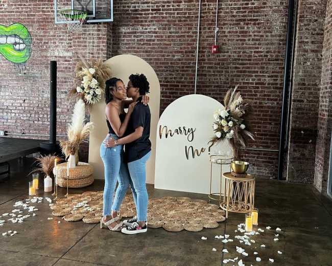 A couple stands together in front of a "Marry Me" display in a venue with exposed brick walls and a basketball hoop.
