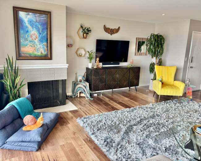 The room features a modern living space with a gray couch, a yellow chair, a television mounted on the wall, and decorative plants.