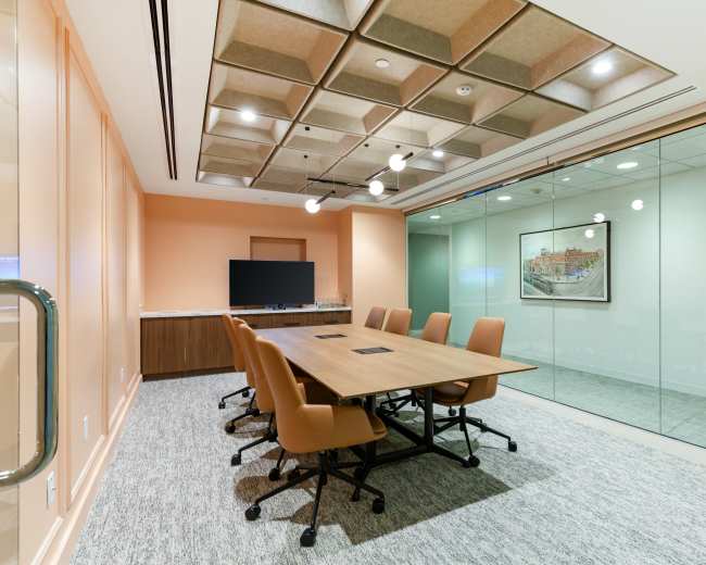 A modern conference room features a large wooden table surrounded by ergonomic chairs, with a wall-mounted screen and a glass wall allowing visibility into the next space.