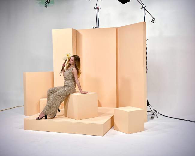 A woman in a patterned jumpsuit poses with flowers on a peach-colored geometric set.