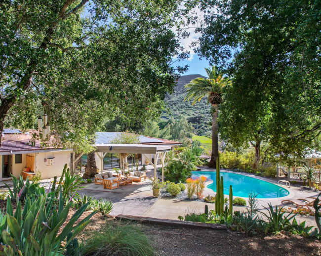 A landscaped outdoor area features a swimming pool surrounded by seating and lush greenery, with a house and a large tree in the background.