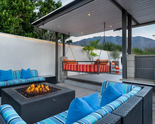 The scene features a modern outdoor seating area with two swinging lounge chairs and a fire pit surrounded by striped cushioned sofas under a covered patio.