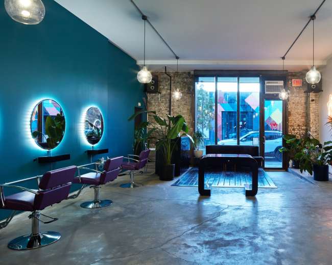 The image shows a modern hair salon with circular mirrors and chairs arranged around the space, featuring plants and large windows that allow natural light to enter.