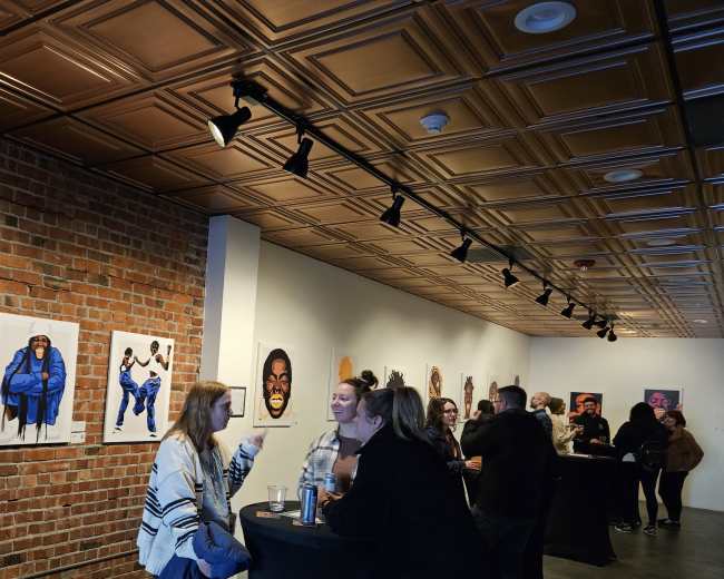 The image shows a gallery event with several people engaging in conversation near tall tables, surrounded by artwork displayed on the walls.