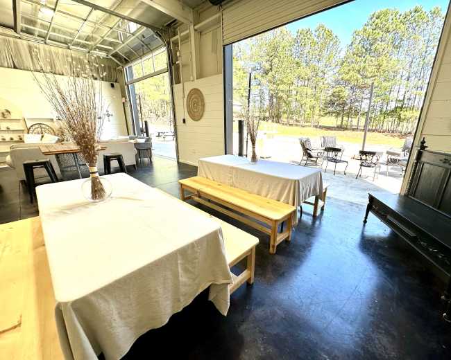 A spacious indoor area with tables covered in white linens, leading to an open door that reveals a view of a tree-lined outdoor space.