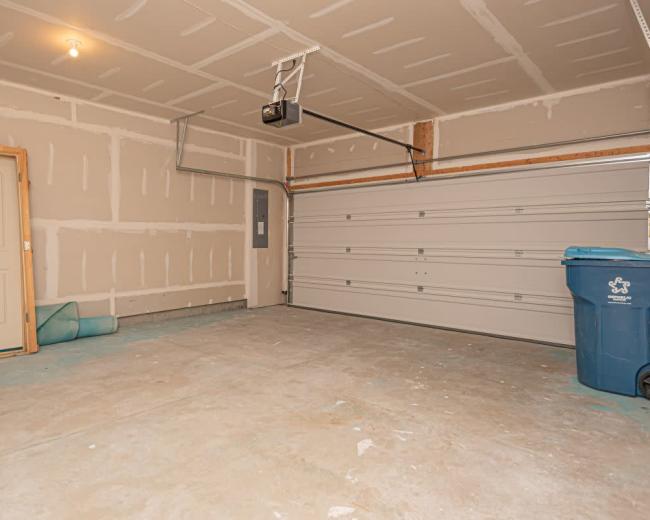 The image shows an empty garage with a concrete floor, unfinished walls, and a blue trash bin in the corner.