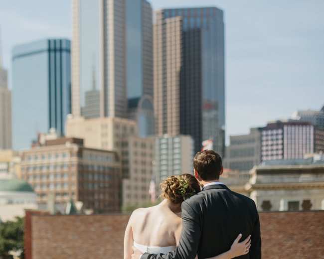 A couple stands together with their backs to the camera, overlooking a skyline of tall buildings in a city.