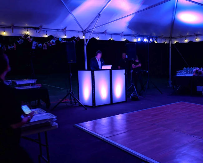 A DJ setup with glowing lights is positioned under a tent, while people socialize and food is displayed on a table.