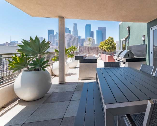 The image shows a spacious balcony with a dining table, grill, and large planters, overlooking a city skyline.