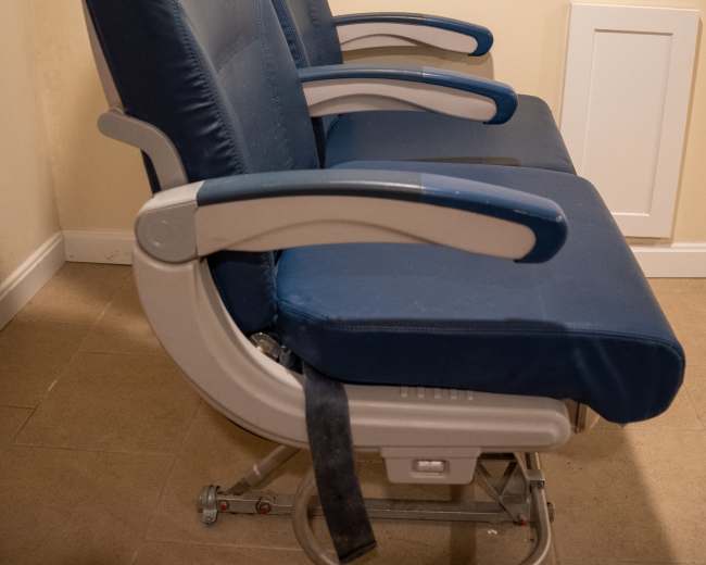 Two blue airplane seats are positioned side by side, accompanied by a window display showing a sky view.