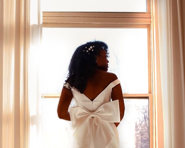 A woman in a flowing white wedding dress with a large bow stands by a window, facing away from the camera.