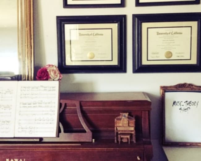 A wooden piano is positioned in front of a wall featuring four framed diplomas and a sheet of music, while a small chair and a whiteboard with text are nearby.