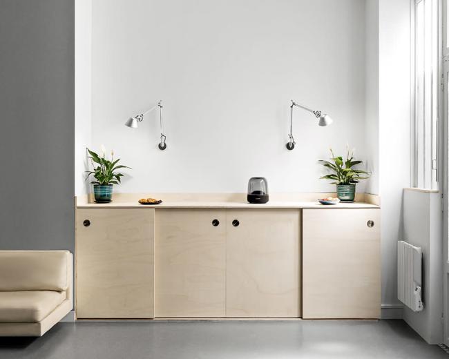 The image shows a minimalist interior with a light wood cabinetry unit, two tabletop plants, and wall-mounted lamps, set against a simple white wall.