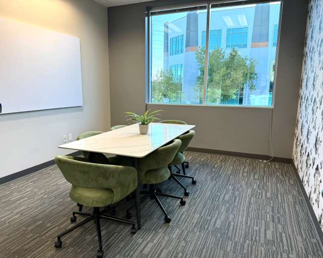 A conference room features a whiteboard, a large table surrounded by green chairs, and windows with a view of a nearby building and trees.