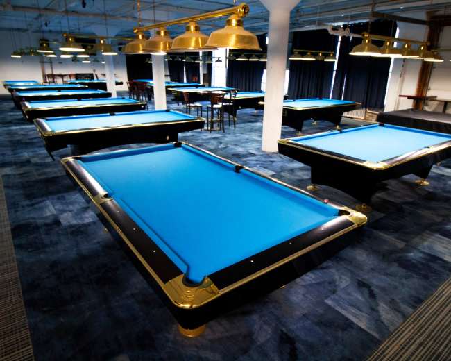 A spacious billiards room with multiple pool tables featuring blue felt tops and industrial-style lighting.