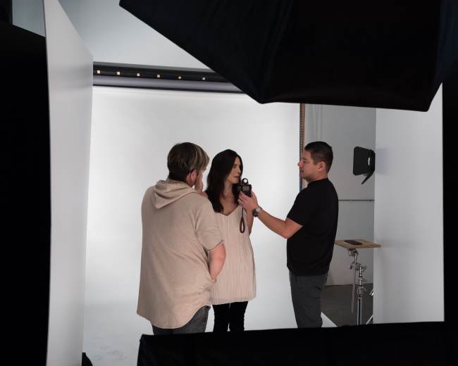 A photographer is adjusting a camera while a model poses, with a stylist discussing in a studio setting.