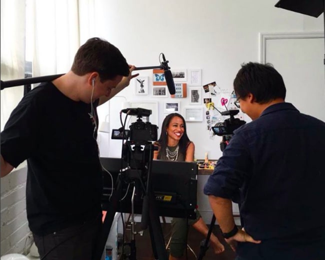 A woman is seated in front of a desk while being filmed by a crew with multiple cameras and microphones.