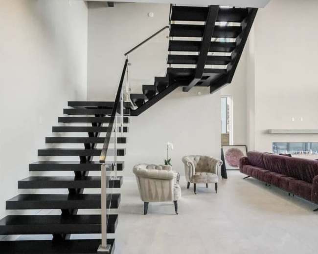 The image shows a modern interior featuring a floating staircase, minimalist furnishings, and large windows providing natural light.