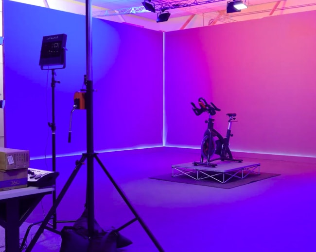 A stationary exercise bike is positioned on a raised platform in a brightly lit studio with purple and blue background lighting.