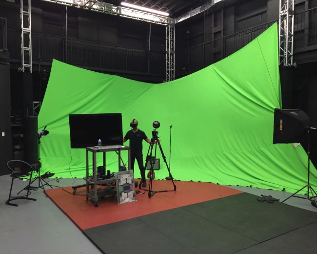 A green screen studio setup features a large green backdrop, a table with equipment, cameras, and lighting in an indoor space.