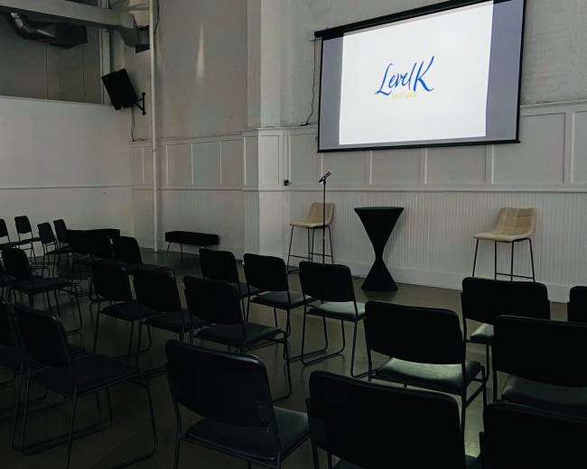 A spacious event room is set up with rows of chairs facing a large screen displaying the text "LevK" on a white background, with two stools and a high table positioned nearby.