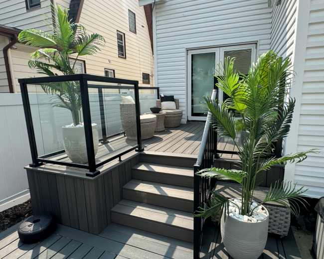 The image shows a wooden deck with stairs, featuring potted plants and a glass railing, adjacent to a house.