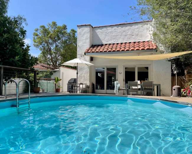 A round swimming pool reflects the clear blue sky next to a modern house with a tiled roof and outdoor furniture.