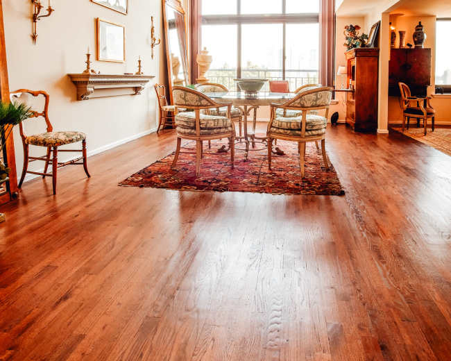 The image shows a spacious, well-lit living area with hardwood floors, a chandelier, and a seating arrangement around a glass table.