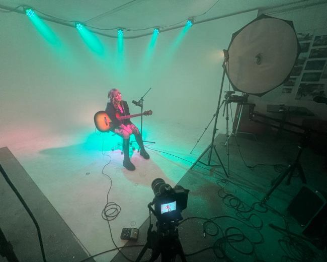 A musician sits with a guitar on a lit stage, surrounded by camera equipment and green lighting.