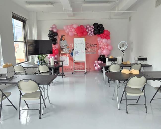 The image shows a bright, spacious room decorated with pink and white balloons, featuring several tables and chairs arranged for an event or workshop.