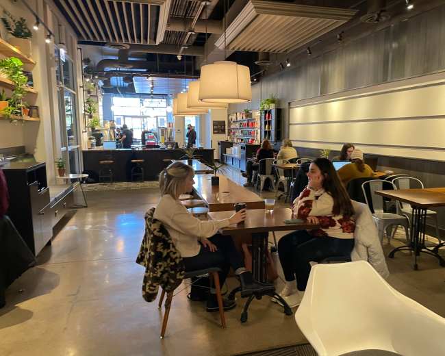 Two women sit at a wooden table in a café, while other patrons are seated at various tables in the background.
