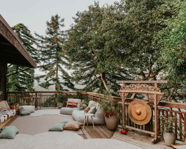 A wooden deck features a cozy seating area with large cushions, potted plants, and decorative elements set against a backdrop of tall trees.