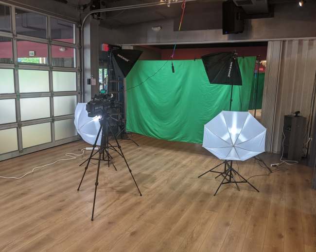 The image shows a studio setup with two softbox lights, a camera on a tripod, and a green screen in a room with wooden flooring.