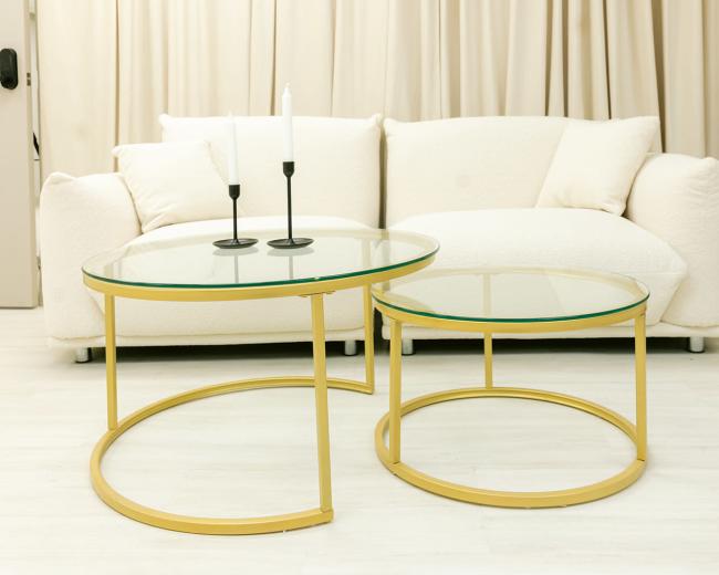 A white couch is positioned against a backdrop of beige curtains, accompanied by two circular glass-top coffee tables with gold frames and a black planter nearby.