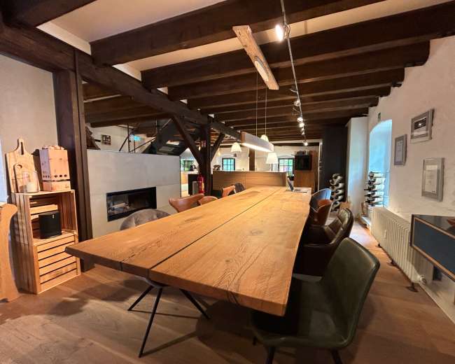 The image shows a spacious dining area with a large wooden table, wooden beams in the ceiling, and modern decor elements.