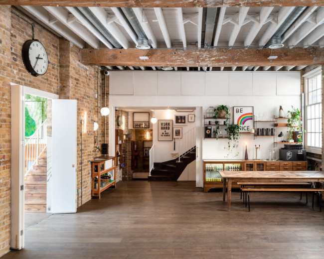The image shows a spacious, modern interior with exposed brick walls, wooden beams, and a long table in a well-lit area featuring various decor and plants.
