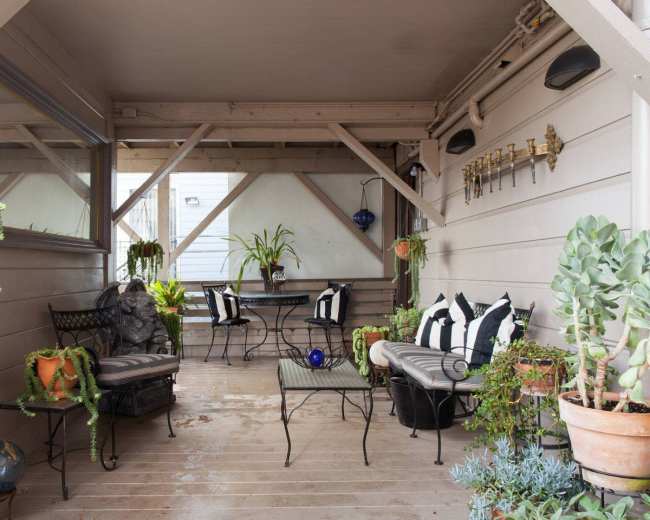 The image shows a covered patio with a table and chairs surrounded by potted plants and decorative lighting.