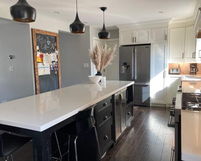 A modern kitchen features a large white island with black chairs, stainless steel appliances, and a vase of decorative grasses.