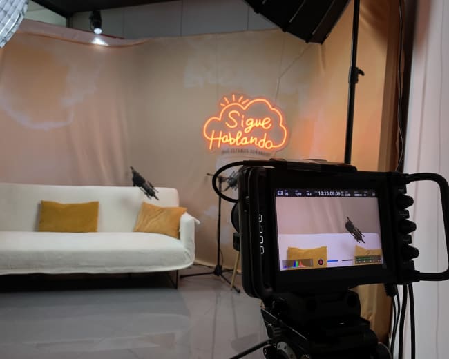 A camera setup is focused on a white sofa with yellow cushions in front of a backdrop featuring a neon sign that reads "Sigue Hablando."