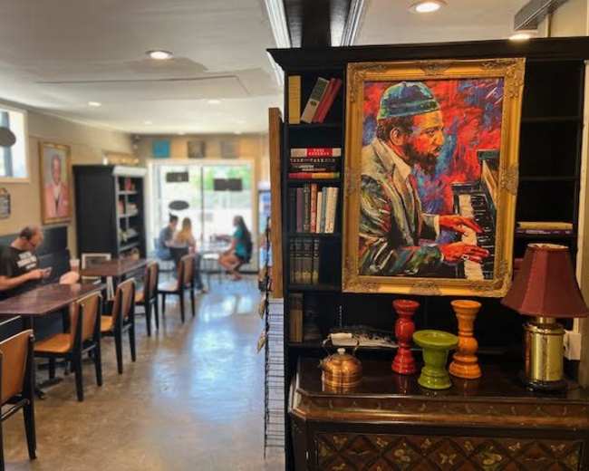 A cozy café interior with a portrait of a pianist on the wall and patrons seated at tables, some engaging with laptops and others in conversation.