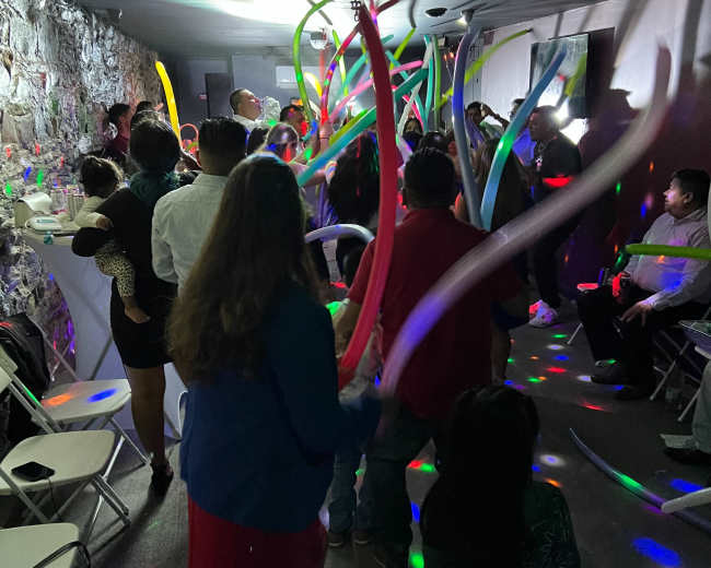 A group of people is dancing in a dimly lit room with colorful lights and long balloons swirling above them.