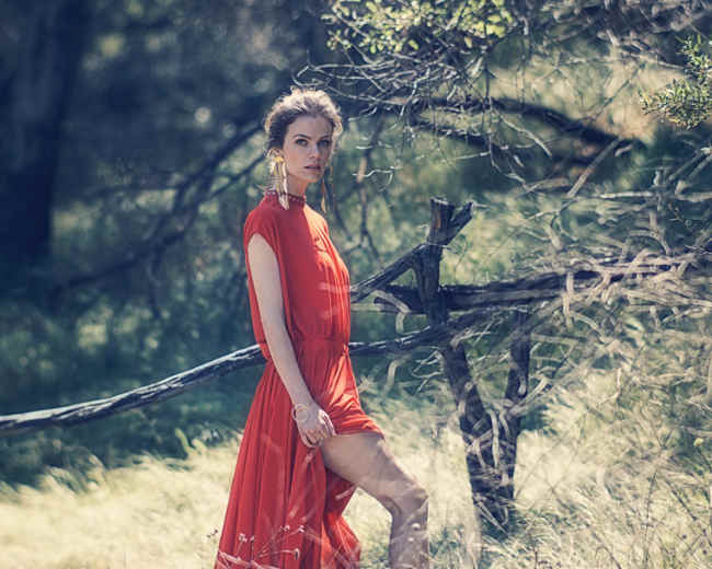 A woman in a red dress stands among tall grass and trees in a natural setting.