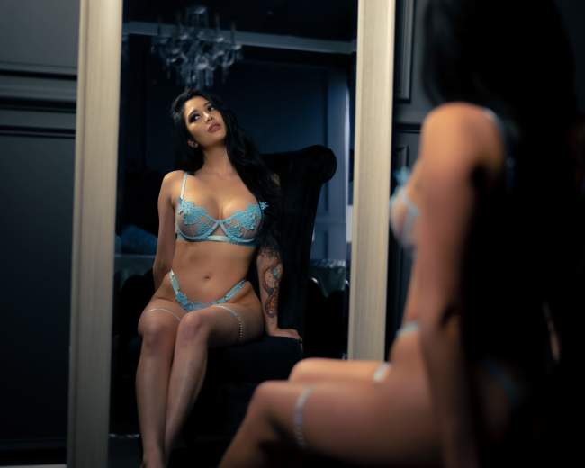 A woman in blue lingerie sits in a chair while looking at her reflection in a mirror.