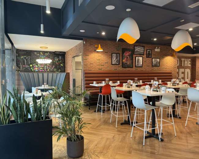 The image shows a modern restaurant interior featuring white tables, colorful chairs, and a mural on a brick wall.