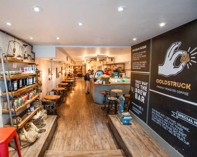 The image shows a modern coffee shop interior with wooden floors, shelves stocked with coffee products, and a counter at the back area.