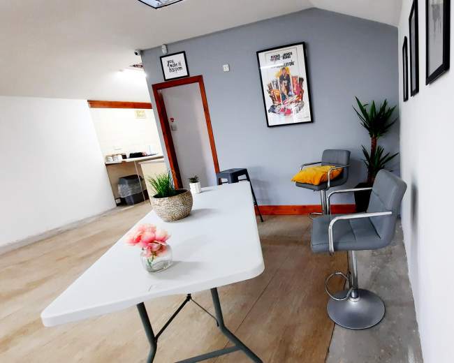 The image shows a room with a table, a bar stool, potted plants, and framed artwork on the walls.