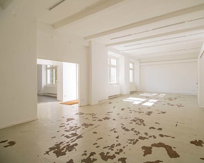 The image shows a spacious, empty room with white walls and a partially worn floor, featuring large windows that allow natural light to enter.