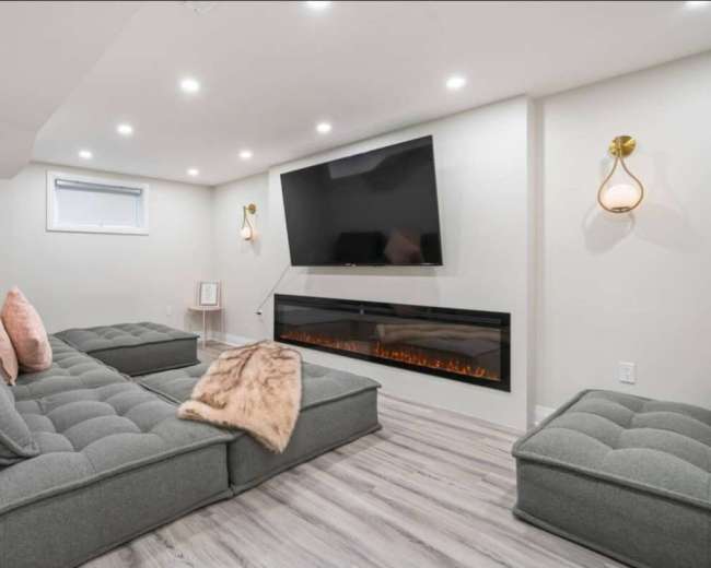 The image shows a modern living room with a large flat-screen TV mounted on the wall, a gray sectional sofa, and an electric fireplace beneath it.